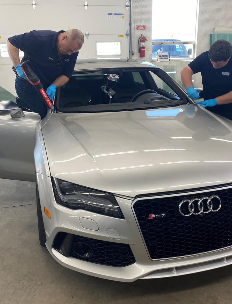 Two men prepping a windshield on a silver Audi in an auto shop to be replaced